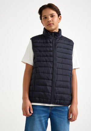 Teenager iført en sort quiltet ærmeløs vest over en hvid kortærmet skjorte og blå jeans, stående mod en ensfarvet baggrund.