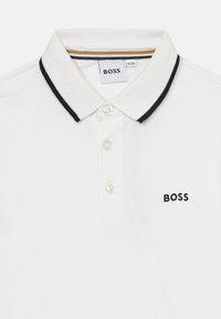 Balta polo marškinėliai, pagaminti iš tekstūruoto audinio, su juodomis juostelėmis apykaklėje, trimis mygtukais ir mažuoju "BOSS" logotipu priekyje.