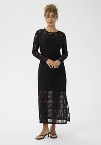 Rochie neagră din dantelă cu mâneci lungi, cu motive complicate și tiv transparent în partea de jos, asociată cu sandale negre cu bretele. Textură netedă.