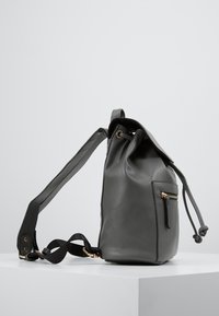 Sac à dos en cuir synthétique gris avec fermeture à cordon, poche latérale zippée et bretelles noires réglables. Texture lisse, design minimaliste.