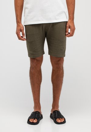 Mann trägt olivgrüne, strukturierte Shorts, ein weißes T-Shirt und schwarze Slip-On-Sandalen und steht vor einem schlichten Hintergrund.