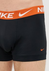 Μαύροι αθλητικοί μποξέρ με πορτοκαλί ελαστική ζώνη που φέρει λευκό κείμενο και λογότυπο "NIKE". Λείος υφασμάτινος τύπος, εφαρμοστή σχεδίαση.