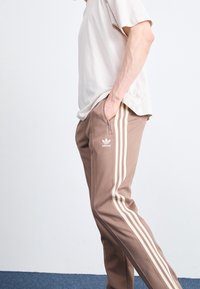 Pantaloni sportivi marroni con tre strisce bianche ai lati, dotati di una tasca con zip e un logo ricamato. Materiale misto cotone, vestibilità comoda.