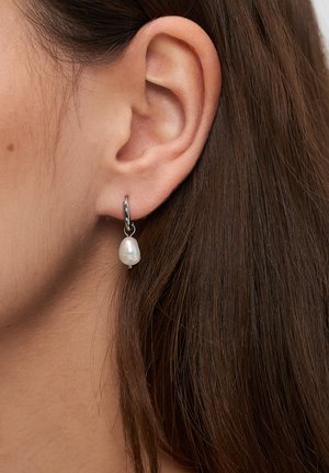 Silberner Creolen-Ohrring mit einer hängenden, unregelmäßig geformten weißen Perle. Nahaufnahme, die die Struktur des Ohrrings und die glänzende Metalloberfläche hervorhebt.