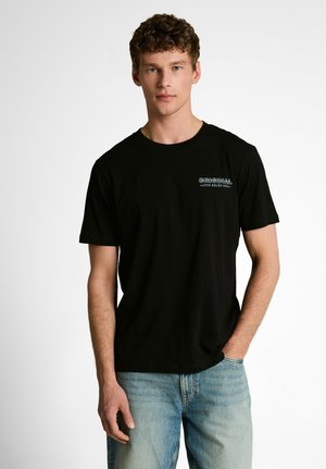 Junger Mann mit lockigem braunem Haar, der ein schwarzes T-Shirt mit dem Text „ORIGINAL 20/07“ und hellblaue Jeans trägt, steht mit einer Hand in der Tasche.