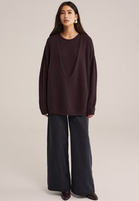 Üks suur burgundia värvi kampsun laia V-kaelusega, madalate õlgade ja pehme tekstuuriga; kombineeritud laiade mustade pükste ja pruunide kingadega.