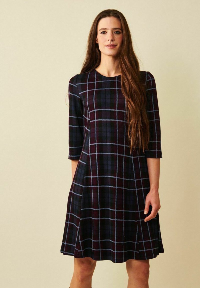Kvinna med långt brunt hår som bär en knälång mörk rutig klänning med trekvartsärmar, stående mot en beige bakgrund.