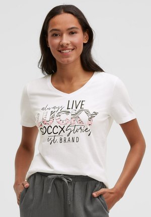 Young woman smiling wearing a white V-neck T-shirt with "always live lucky" text and gray drawstring pants, hands in pockets.