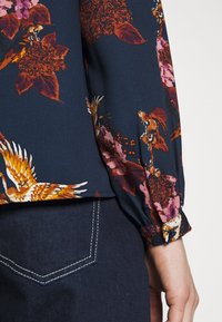Blusa navy con motivo floreale nei toni dell'arancione e del rosa. Presenta maniche arricciate e polsini con bottoni. Abbinata a jeans scuri.