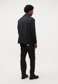 Zwarte enkel-breasted jas met brede revers, gecombineerd met een donkere corduroy broek. Gladde stoffen afwerking, getoond vanuit een achteraanzicht.