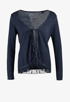 Marineblå cardigan med flæsekant, båret over en hvid top med blonder, kombineret med blå revnede jeans og sorte støvler med høje hæle, bærende en sort taske.