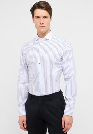 Ung mand iført en hvid skjorte med tynde lodrette striber og sorte bukser, stående mod en ensartet lys baggrund.