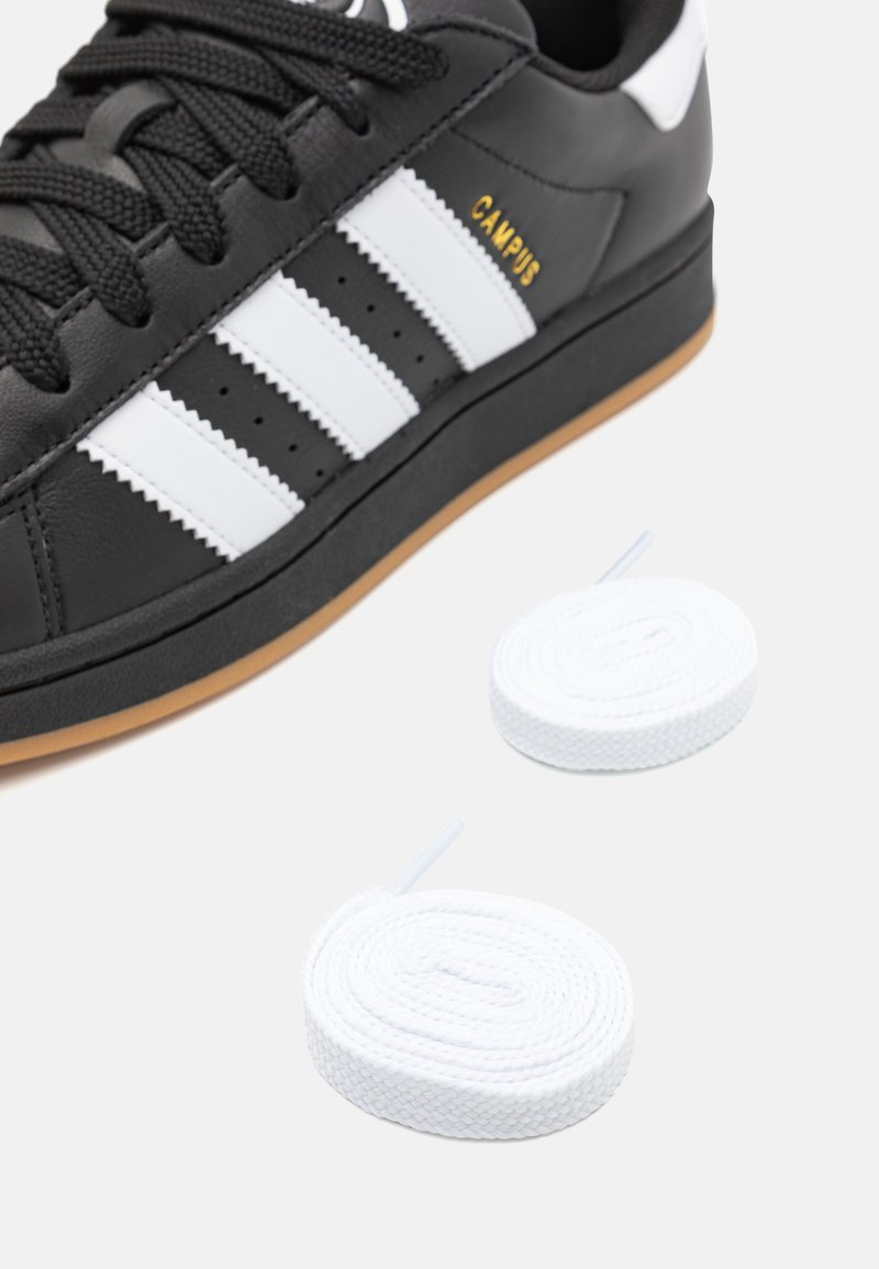 Zapatilla de cuero negra con rayas blancas, suela de goma y letras doradas. Incluye dos agujetas planas blancas. Punta redondeada y parte superior texturizada.