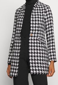 Manteau à motif pied-de-poule en noir et blanc, manches longues, col cranté, fermeture à un bouton et tissu texturé.