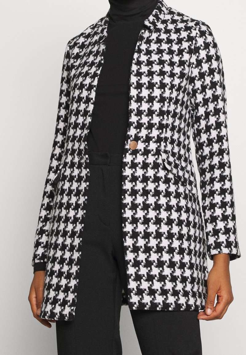 Manteau à motif pied-de-poule en noir et blanc, manches longues, col cranté, fermeture à un bouton et tissu texturé.