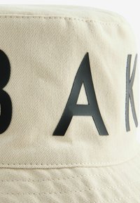 Beige Bucket Hat aus Canvas, mit großen schwarzen Buchstaben "B A K" auf der Vorderseite geprägt. Die Nähte sind entlang des Schirms sichtbar.
