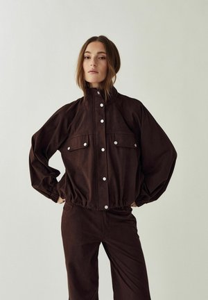 Žena v tmavohnedej zapínanej bunde s nadýchanými rukávmi a nohaviciach v rovnakom odtieni stojaca s rukami v bok na jednoduchom pozadí.