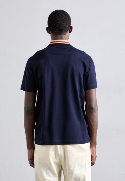 Homme debout de dos, portant une chemise à manches courtes bleu marine avec un col jaune et rouge, et un pantalon crème, contre un fond uni.