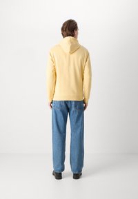 Un homme se tenant de dos, portant un sweat à capuche jaune clair, un jean bleu et des chaussures noires, sur fond blanc uni.