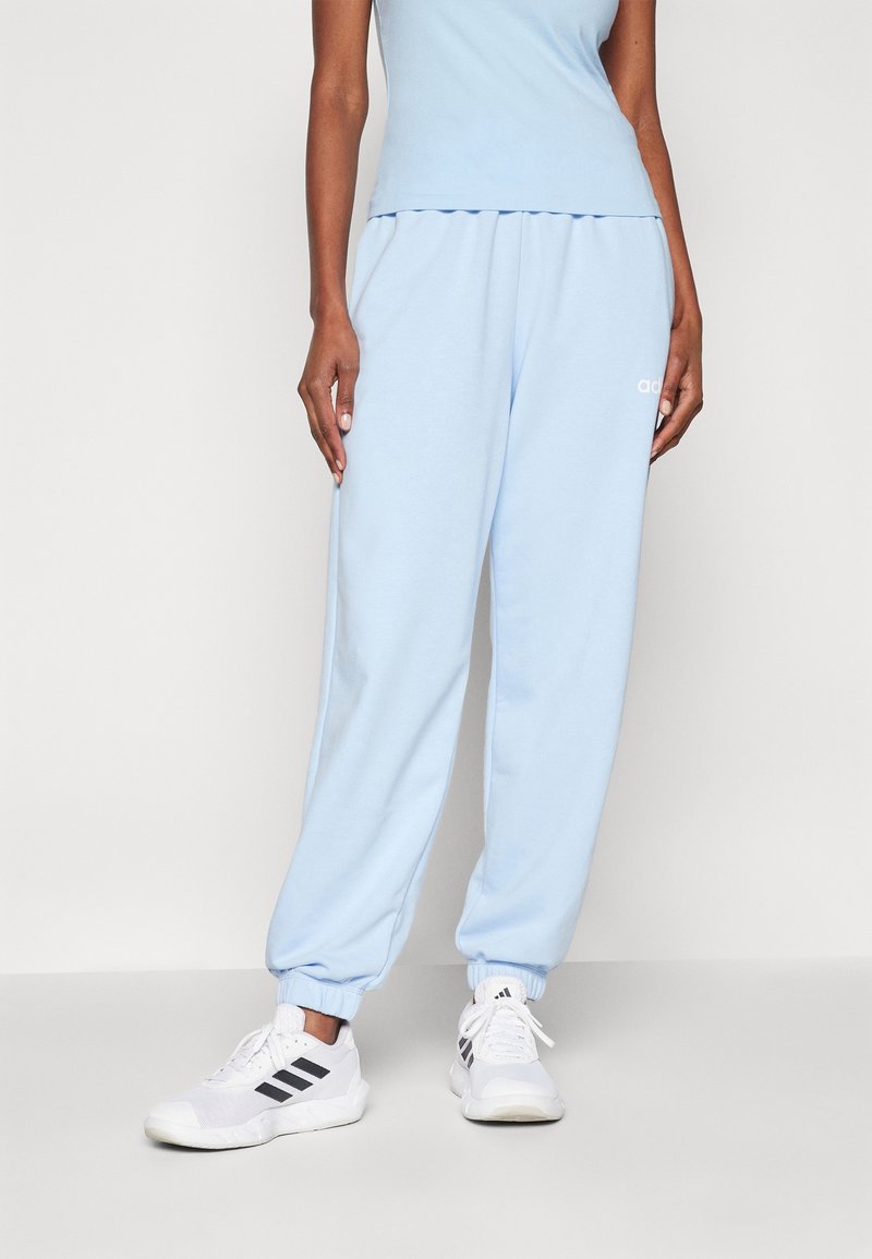 Person som bär ljusblå mjukisbyxor och vita Adidas-sneakers med svarta ränder, stående mot en enkel ljus bakgrund.