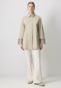 Chemise beige à boutonnières avec des manches longues et transparentes. Associée à un pantalon évasé blanc et des chaussures de couleur claire. Design minimaliste, texture lisse.