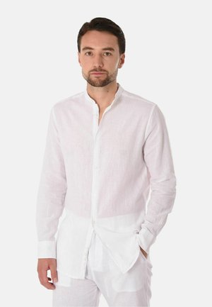 Uomo con capelli corti e scuri e barba, che indossa una camicia bianca a maniche lunghe abbottonata e pantaloni bianchi davanti a uno sfondo semplice.