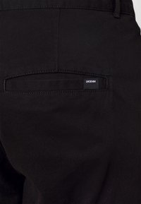 Pantaloni in denim nero con una tasca posteriore che presenta una piccola etichetta "DRODENIM". Il tessuto è liscio con una leggera lucentezza.