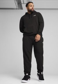 Hoodie noir avec une poche frontale et un logo, associé à un pantalon de jogging assorti, avec des poignets élastiques et des baskets noires avec des accents roses.