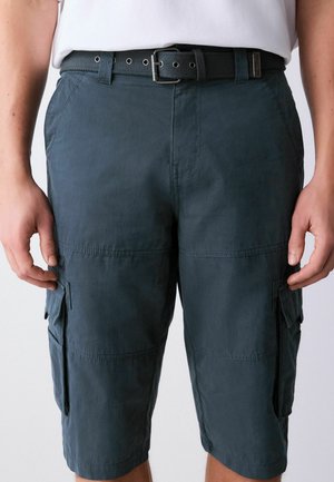 Mężczyzna noszący ciemnoniebieskie szorty typu cargo z bocznymi kieszeniami i czarnym plecionym paskiem, ręce swobodnie opuszczone wzdłuż ciała.