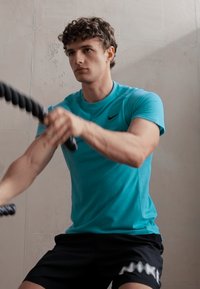 Türkisfarbiges Sportshirt mit kurzen Ärmeln, kombiniert mit schwarzen Shorts mit einem weißen Muster. Person verwendet Fitnessseile in einem Innenraum.