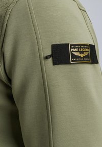 Maslinasto zelena tkanina s crnim pravokutnim izrezom na kojem je zlatni natpis "PME Legend" i krilati emblem na rukavu.