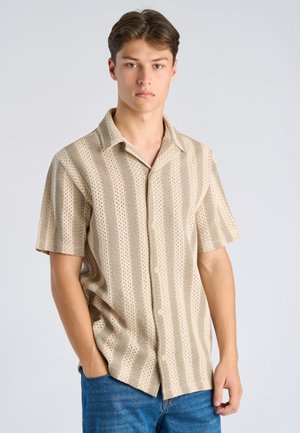 Junger Mann trägt beige, kurzärmliges Hemd mit Knöpfen und vertikalen Streifen sowie blaue Jeans, steht mit einer Hand in der Tasche vor grauem Hintergrund.