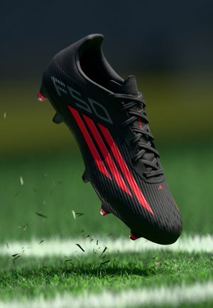 Schwarzer Fußballschuh mit roten Streifen, strukturiertem Obermaterial und Schnürsenkeln. Verfügt über leuchtend rote Studs und wird über Gras mit Schmutz dargestellt.