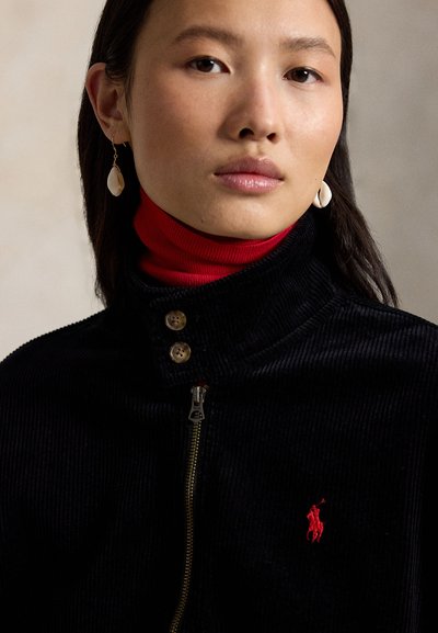 Νεαρή γυναίκα φοράει μαύρο ζακέτα με φερμουάρ και κόκκινο λογότυπο, κόκκινο ζιβάγκο από μέσα, και λευκά κρεμαστά σκουλαρίκια πάνω σε ουδέτερο φόντο.