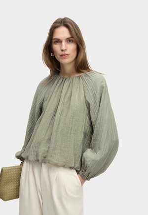 Blouse plissée vert clair avec de longues manches bouffantes et un ourlet élastiqué. Associée à un pantalon couleur crème et un petit sac texturé.