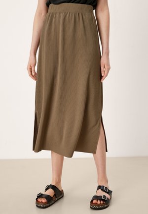 Femme portant une jupe midi côtelée vert olive avec des fentes latérales et des sandales noires à boucle, debout sur un sol beige contre un mur clair.
