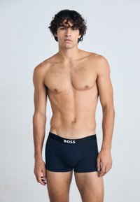 Schwarze Boxer-Briiefs mit einem weißen Logobund, aus dehnbarem Material und einem figurbetonten Design, getragen von einem männlichen Modell mit kurzen Haaren.