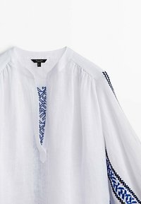 Witte linnen blouse met een V-hals en borduurwerk in blauw en zwart langs de mouwen. Beschikt over licht opbollende mouwen en een gestructureerde stof.