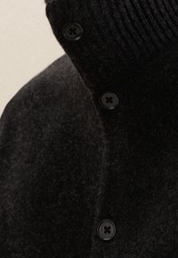 Musta villatakki, jossa on korkea kaulus, poimuttainen pinta ja suuret pyöreät napit edessä. Pehmeä, hieman pörröinen pinta.