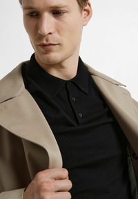 Svart polo-skjorte med tre knapper, under en beige jakke. Nærbilde viser stofftekstur og kragedetaljer. Nøytral bakgrunn.