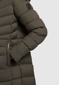 Olive green padded coat featuring quilted texture, a black logo patch on the sleeve, and a zippered side pocket.