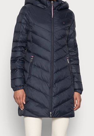 Femme portant un long manteau d'hiver matelassé bleu marine avec une capuche, une fermeture éclair devant, des poches zippées et un pantalon blanc.