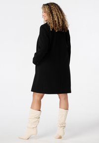 Femme aux cheveux bouclés portant un manteau noir et des bottes à talons crèmes, se tenant dans une pose de trois-quarts de dos sur fond blanc.