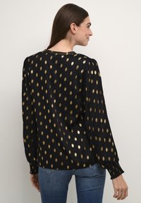 Blusa nera con motivo a pois dorati, maniche lunghe a sbuffo, polsini arricciati e scollatura arrotondata. Tessuto liscio con una leggera lucentezza.