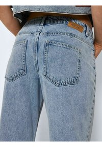 Jeans de ganga azul claro com um corte alto e perna larga. Apresenta dois bolsos traseiros e um pequeno rótulo castanho na cintura.