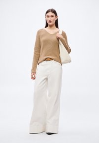 Beige strikkegenser med V-hals, kombinert med hvite vide bukser. Tilbehør inkluderer et brunt belte og en beige skulderveske.