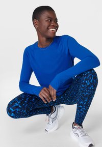 Langsleeve blauwe sporttop gecombineerd met gestreepte zwart-blauwe leggings, aangevuld met witte sneakers met accenten.