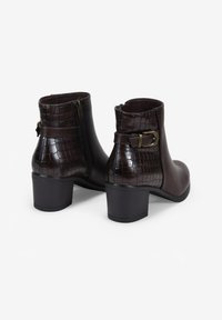 Botas de tobillo marrones oscuras con tacones de bloque, textura de cocodrilo en la parte trasera y lateral, cremallera lateral y correas con hebilla ajustable.