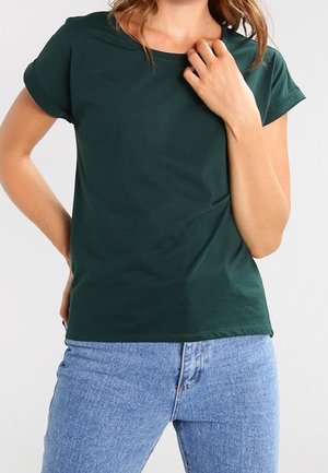 Kobieta ubrana w ciemnozielony krótki rękaw t-shirt i niebieskie jeansy, jedną ręką dotyka kołnierza, a drugą talii.