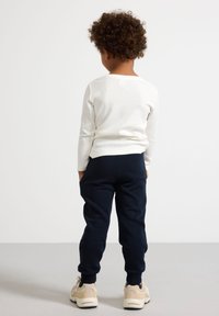 Weißes langärmeliges geripptes Shirt, marineblaue Jogginghose mit elastischen Bündchen und hellfarbige Sneakers. Der Rücken des Kindes zeigt zur Kamera.